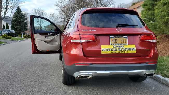 An open car door on a red SUV demonstrating how dooring accidents happen when a driver or passenger suddenly opens a car door directly in front of a passing motorcycle or bicycle in traffic. The SUV has a HURT 911® license plate and a Be Aware Motorcycles Are Everywhere® car sign on the back.
